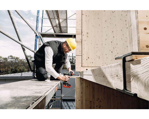 Ein Mann mit Schutzhelm arbeitet mit einem Akku-Bohrschrauber auf einem Gerüst an einer Holzkonstruktion.