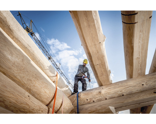 Un homme travaille avec une perceuse sans fil sur la construction d''une maison en bois, avec une grue de chantier en arrière-plan.