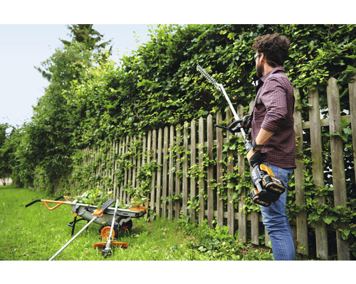 Ein Mann schneidet eine Hecke mit einer Akku-Heckenschere im Garten. Im Hintergrund sind eine Schubkarre und ein Rasentrimmer zu sehen.