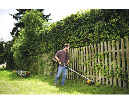 Ein Mann trimmt Gras mit einem Akku-Rasentrimmer in einem Garten mit Holzzaun und Hecke.