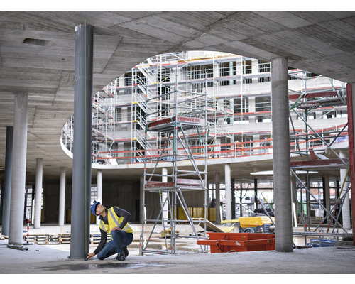 Un ouvrier du bâtiment prend des mesures sur une colonne en acier sur un chantier, à l''arrière-plan un gros œuvre avec un échafaudage.