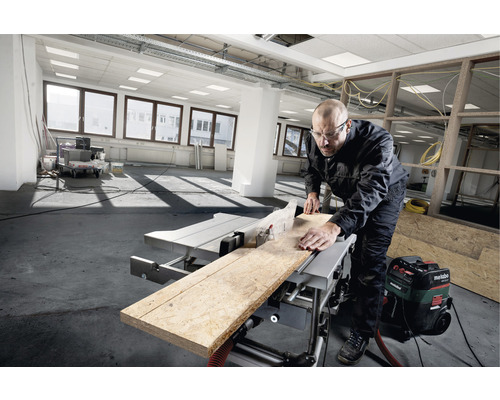Ein Mann benutzt eine Tischkreissäge, um ein Holzbrett zu schneiden. Im Hintergrund ist ein Staubsauger zu sehen.