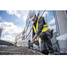 Un ouvrier du bâtiment est agenouillé et utilise une visseuse sans fil sur un chantier.