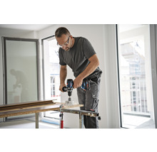 Un homme perce un trou dans une planche de bois avec une perceuse sans fil. Il porte des lunettes de protection et des vêtements de travail.