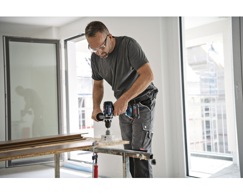 Ein Mann bohrt mit einer Akku-Bohrmaschine ein Loch in ein Holzbrett. Er trägt eine Schutzbrille und Arbeitskleidung.