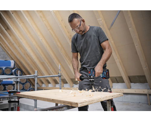 Ein Mann bohrt mit einem Akku-Bohrschrauber ein Loch in ein Holzbrett in einem Dachgeschoss.