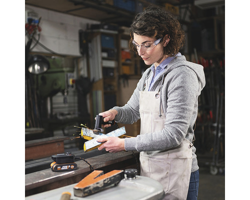 Une femme travaille avec une meuleuse d''angle sans fil dans un atelier, portant des lunettes de protection.