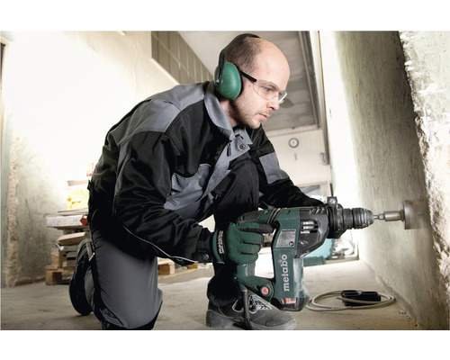 Un homme perce un trou dans un mur en béton avec une perceuse Metabo. Il porte une protection auditive et des lunettes de sécurité.