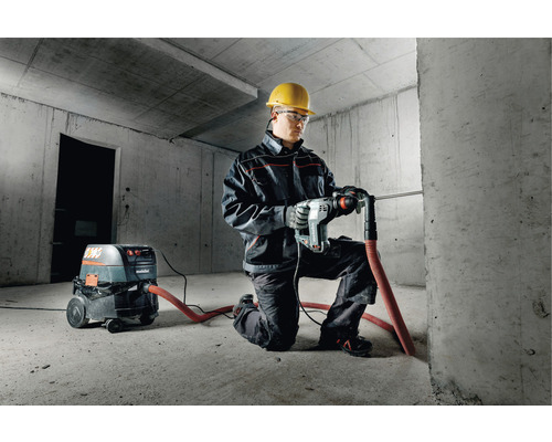 Homme avec casque de protection, lunettes de protection et marteau perforateur Metabo perçant un mur en béton. Un aspirateur de la marque Metabo se trouve à côté.