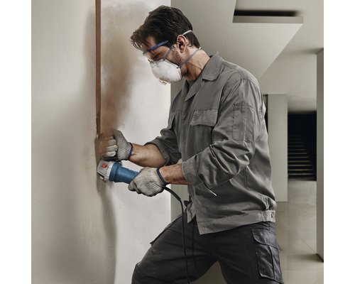 Handwerker schleift eine Wand mit einem Winkelschleifer, während er Schutzbrille und Staubmaske trägt.