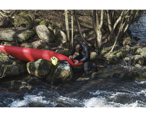 Un homme répare un kayak sur une rive avec des rochers et des arbres.