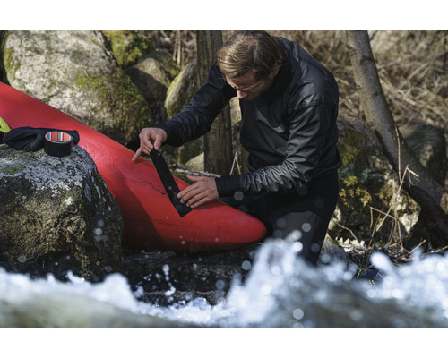 Un homme répare un kayak rouge avec du ruban adhésif noir près des rochers et de l''eau courante.