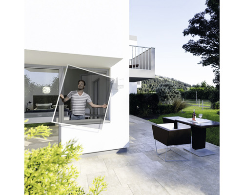 Un homme installe un cadre de protection contre les insectes sur une fenêtre d''une maison moderne. Un jardin avec un coin salon en arrière-plan.