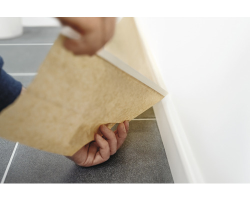 Person verlegt eine Fussleiste mit Papierunterlage entlang einer Wand auf einem gefliesten Boden.
