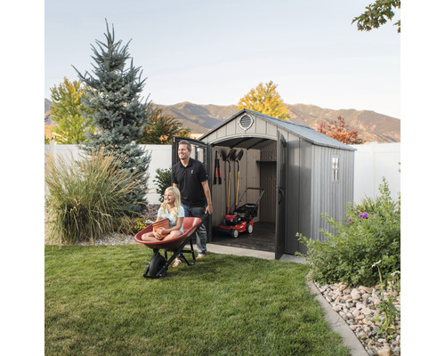 Szene mit einem Geräteschuppen, in dem eine Rasenmähermaschine und Gartengeräte aufbewahrt werden, während ein Mann ein Mädchen in einer Schubkarre schiebt.