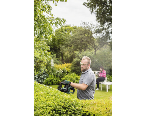 Ein Mann schneidet mit einer Heckenschere eine Hecke in einem Garten. Eine Frau sitzt im Hintergrund.