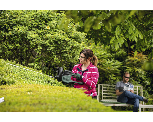 Une femme taille une haie avec un taille-haie dans le jardin, tandis qu''un homme se repose sur un banc.