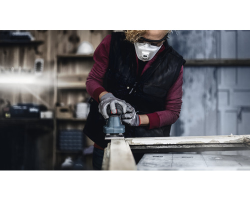 Frau schleift Holz mit einem Exzenterschleifer, getragen werden Schutzbrille, Atemschutzmaske und Handschuhe.
