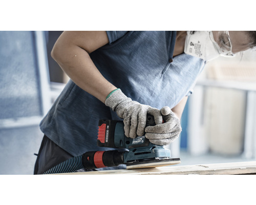 Eine Person schleift Holz mit einem Schwingschleifer, ausgestattet mit Staubabsaugung, während sie Schutzbrille, eine Staubmaske und Handschuhe trägt.