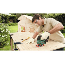 Un homme travaille avec une scie sauteuse sur une planche de bois dans le jardin, avec une maison en bois à l''arrière-plan.