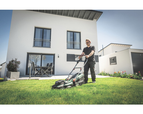 Un homme tond le gazon avec une tondeuse à batterie dans le jardin devant une maison moderne