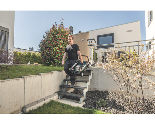 Un homme descend un escalier dans un jardin en portant une tondeuse à gazon pliée