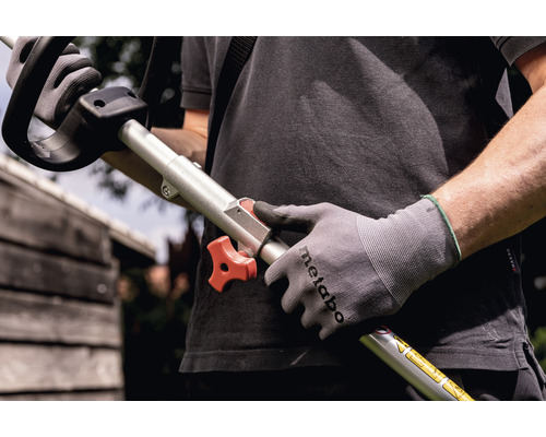 Une personne tient une débroussailleuse avec un bouton rotatif rouge à la main et porte des gants de travail.