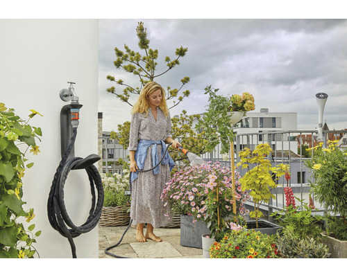 Frau bewässert Pflanzen auf einem Balkon mit einem Gartenschlauch an einer Wandhalterung.