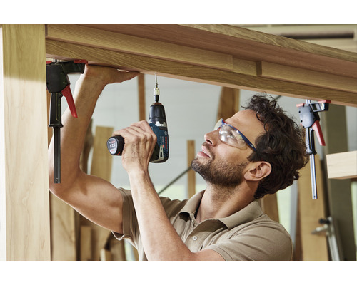 Artisan travaillant avec un tournevis sans fil sur un cadre en bois. Outils et équipements de protection sont disponibles.