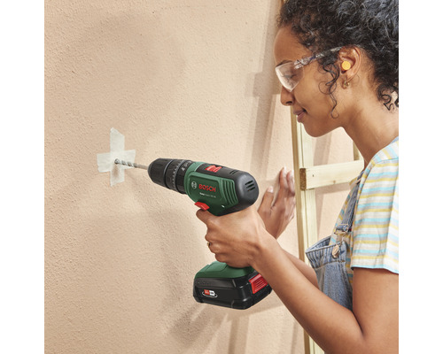Une femme perce un trou dans un mur avec une perceuse sans fil. Elle porte des lunettes de protection.