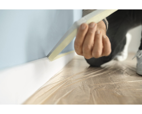 Eine Person klebt Malerkrepp an einer Wand mit Fussleiste, um sie vor Farbspritzern zu schützen.