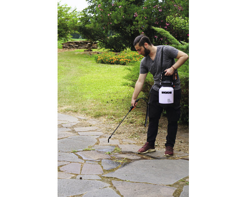 Eine Person benutzt einen Gartensprüher auf einer Steinterrasse zur Unkrautbekämpfung. Im Hintergrund sind Büsche und ein Garten zu sehen.