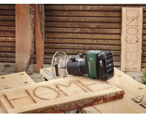 Akku Oberfräse beim Gravieren des Wortes HOME in ein Holzbrett, im Hintergrund ein Brett mit dem Wort WILLKOMMEN
