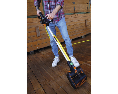 Eine Person reinigt eine Holzterrasse mit einer elektrischen Bürste. Die Bürste hat einen langen Griff und einen Bürstenkopf.