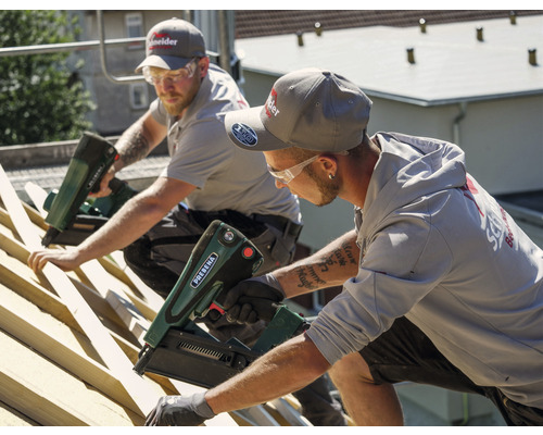 Zwei Dachdecker befestigen mit einem Druckluftnagler Latten auf einem Dach.