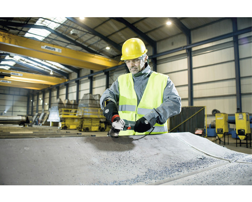 Ein Arbeiter mit Schutzhelm und Schutzweste schleift ein Metallstück mit einer Schleifmaschine in einer Werkshalle.