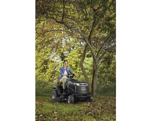 Une femme conduit un tracteur de jardin dans un jardin arboré.