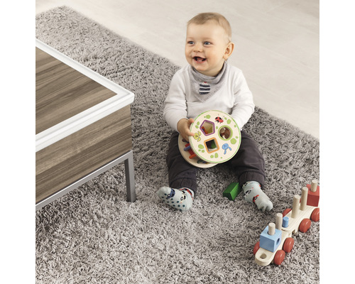 Ein Baby sitzt auf einem grauen Teppich und spielt mit Holzspielzeug, darunter ein Formensortierer und ein Holzzug.