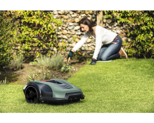 Un robot de tonte est posé sur une pelouse, tandis qu''une femme travaille dans le jardin en arrière-plan.