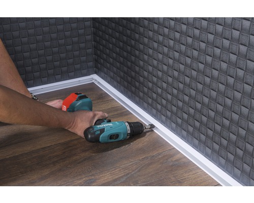 Handwerker montiert eine Sockelleiste mit einem Akku-Bohrschrauber in einem Raum mit Holzboden und strukturierter Wand