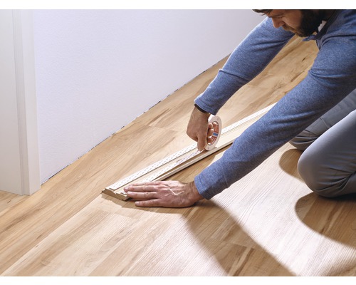 Eine Person befestigt eine Sockelleiste mit einem Klebeband auf einem Holzboden.