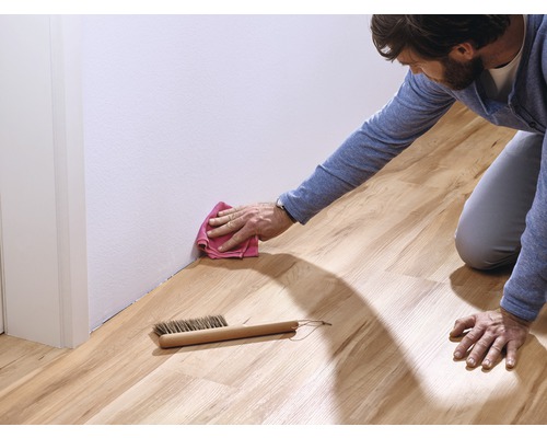Mann reinigt Laminatboden mit einem Tuch und einer Bürste entlang einer weißen Wand