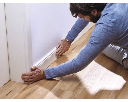 Ein Mann montiert eine weiße Sockelleiste an einer Wand in einem Zimmer mit Holzboden.