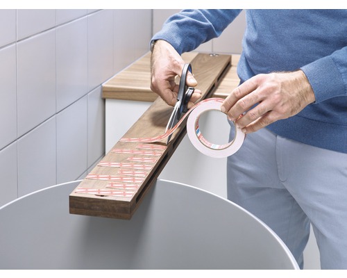 Une personne coupe du ruban adhésif double face avec des ciseaux sur une planche en bois.