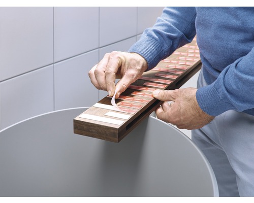 Une personne retire le film protecteur d''une bande adhésive sur une latte en bois au-dessus d''une poubelle.