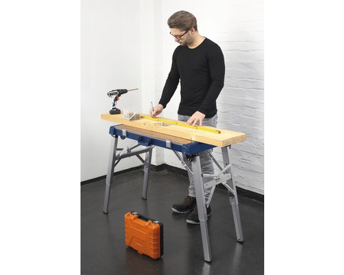 Un homme travaille sur une planche de bois avec un établi, une visseuse sans fil, des vis et un mètre ruban.