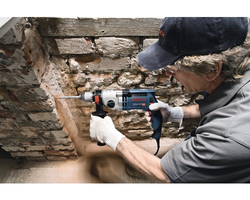 Mann bohrt mit einer Schlagbohrmaschine ein Loch in eine Ziegelwand. Bosch Logo.
