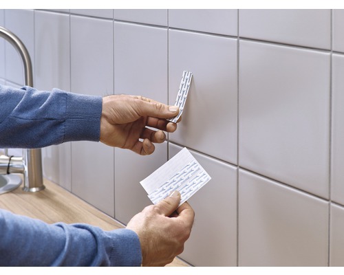 Une personne fixe des bandes adhésives double face sur un mur carrelé.