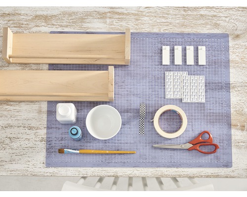 Deux étagères en bois, de la peinture, un pinceau, des ciseaux, du ruban adhésif et du papier décoratif sont posés sur un tapis de découpe.