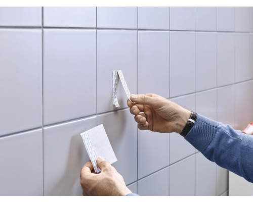 Un homme fixe un objet sur un mur carrelé avec du ruban adhésif double face.